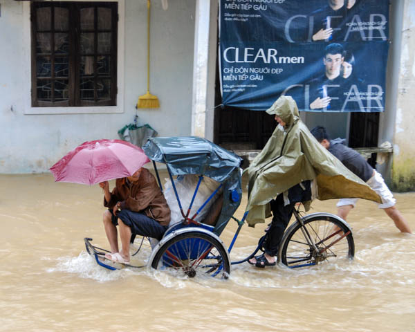 Rikschafahrer und Gast mit Regenschirm fahren durch das Hochwasser